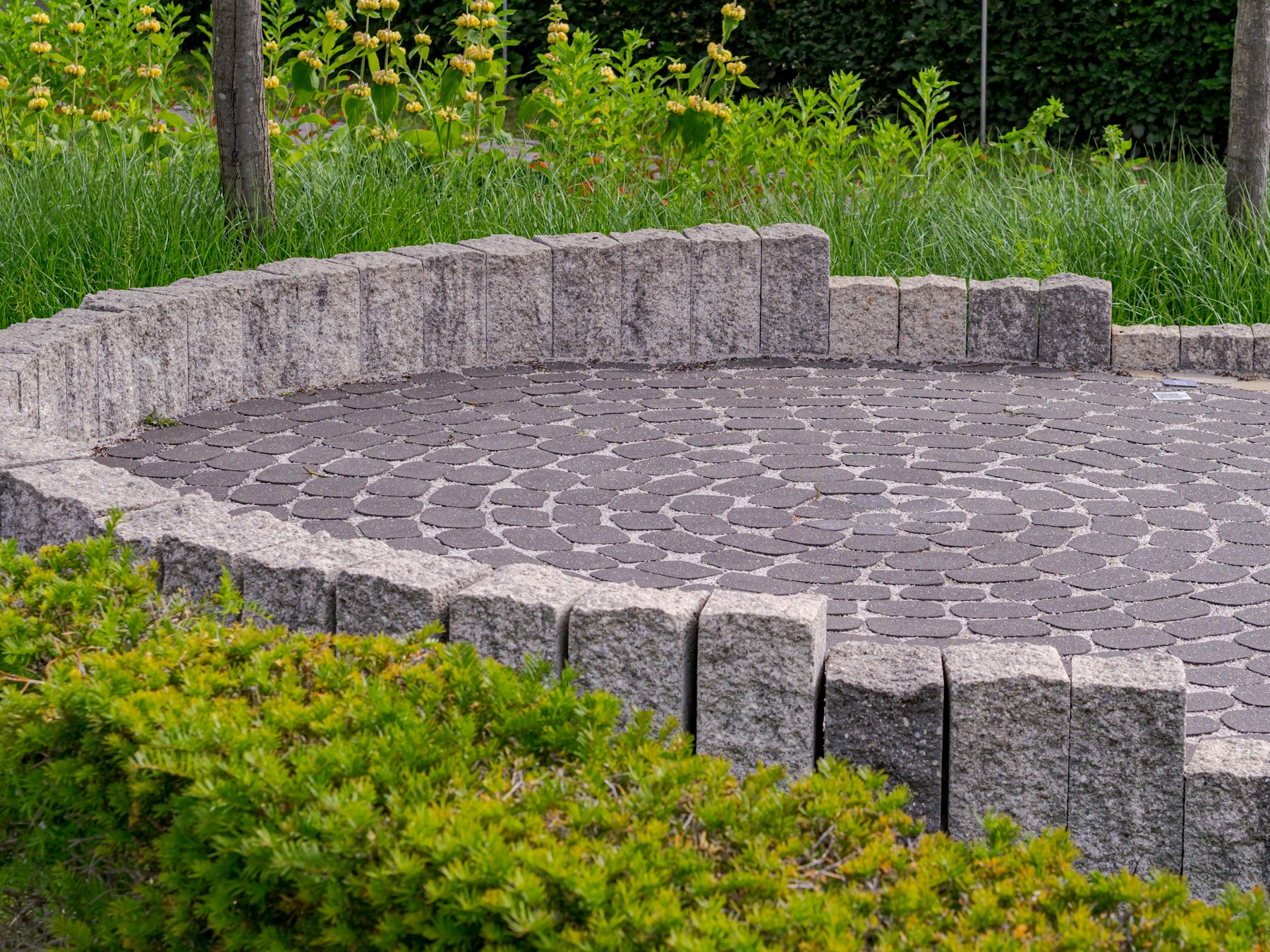 Circular paver patio in a fan pattern, edged with a low stone palisade wall and surrounded by green plantings