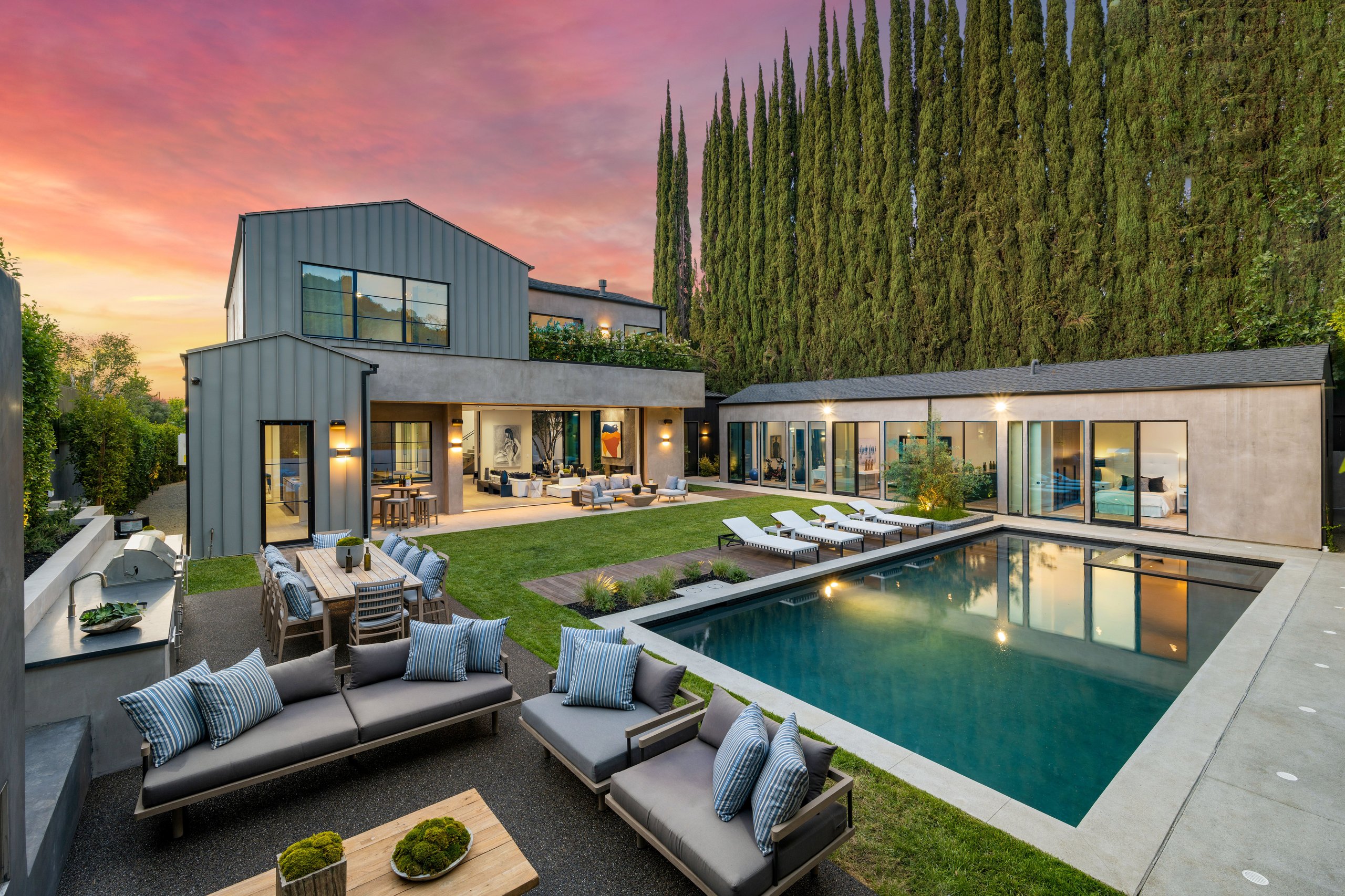 Modern home with pool, lounge chairs, and outdoor dining framed by cypress trees at dusk